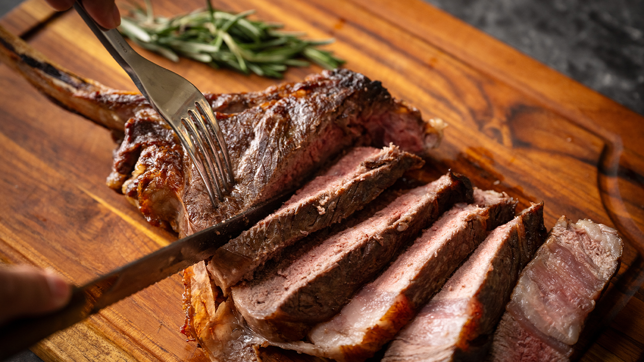 Sliced medium-rare steak being carved on a wooden board with herbs.