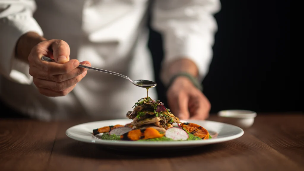 Chef drizzling sauce over a plated gourmet dish in a restaurant kitchen.