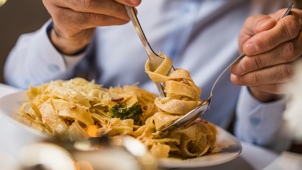 Close-up of a person twirling creamy pasta using a fork and spoon at an Italian restaurant.