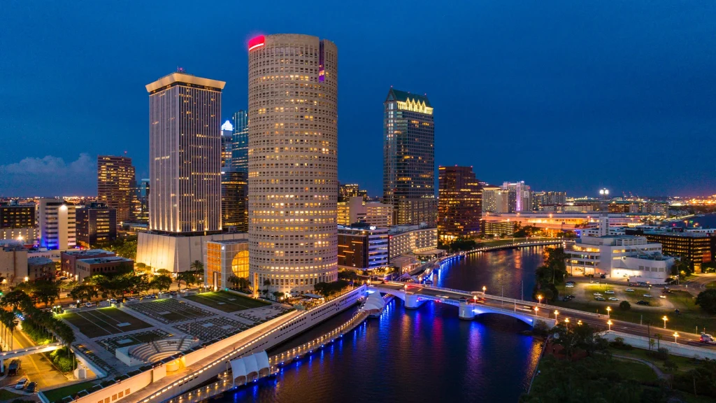 Night skyline of downtown Tampa with illuminated Riverwalk and bridges along the Hillsborough River.