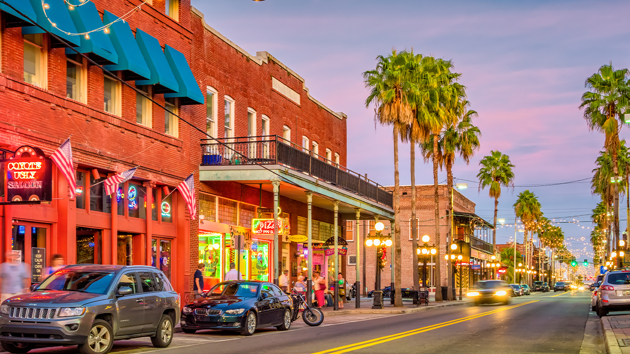 Colorful street view of historic Ybor City with neon signs, restaurants, and palm trees at dusk.