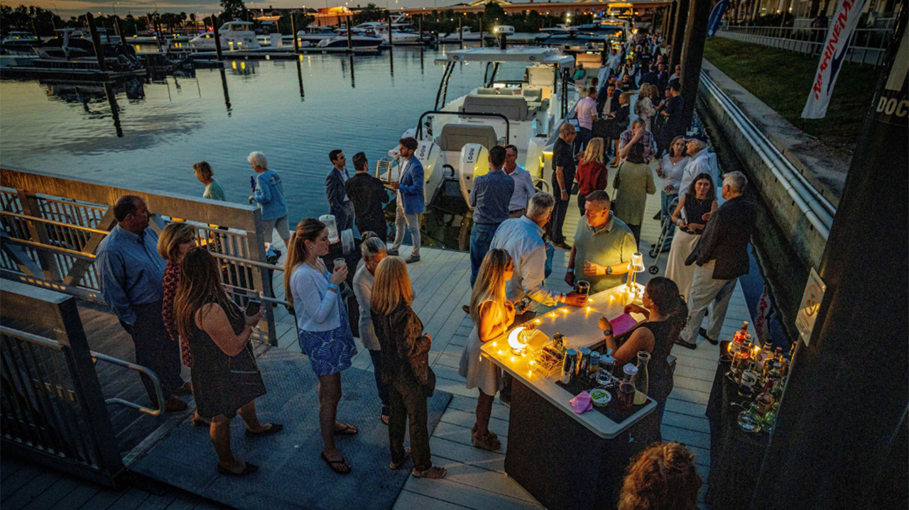 Evening gathering on a dock beside moored boats with people standing near a lit table.