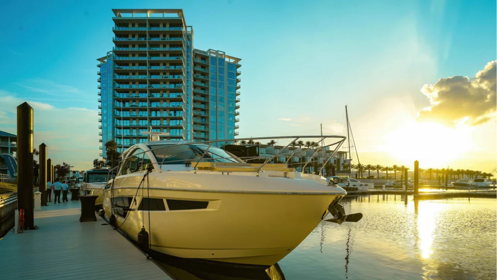 Large luxury yacht docked at a waterfront marina with a modern high-rise building and sunset reflecting on the water.