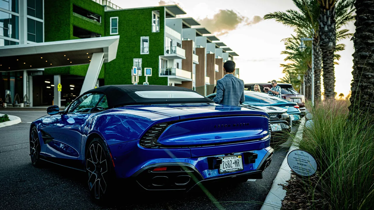 Blue sports car parked near modern buildings and palm trees at dusk.