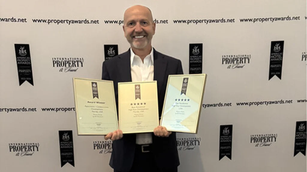 Executive director holding three Americas Property Awards in front of International Property Awards backdrop
