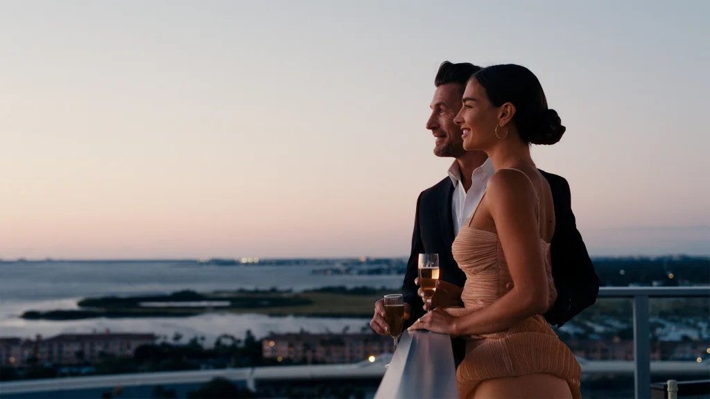 Well-dressed couple holding champagne on rooftop overlooking water at dusk.